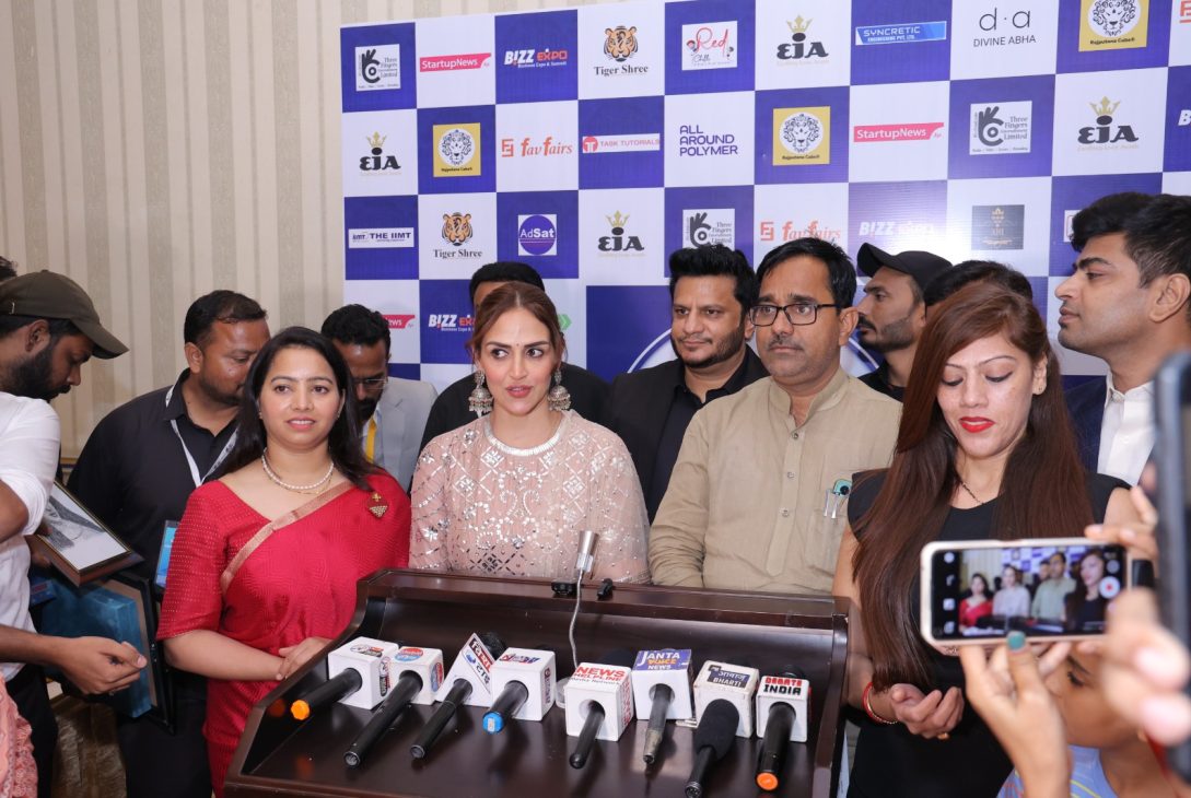 A group of people standing in front of a backdrop with various logos, giving a press conference. Several microphones from different news agencies are placed on a podium in front of them at Bizz Expo & Summit.